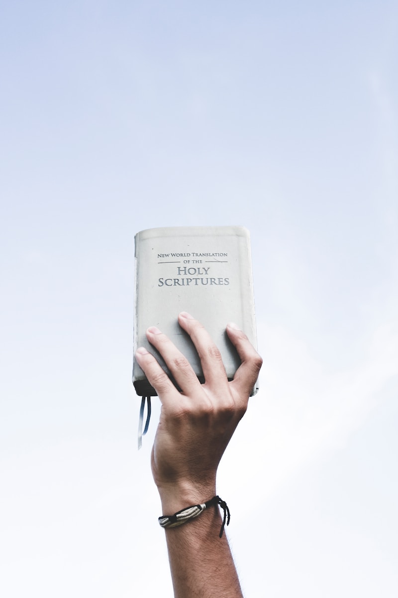 A hand holds up a Bible titled "New World Translation of the Holy Scriptures" against a clear sky, symbolizing faith and devotion.