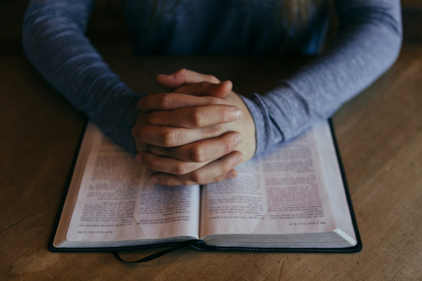 Hands clasped in prayer over an open book on a wooden table, suggesting contemplation and devotion. This image reflects themes of spirituality or study.