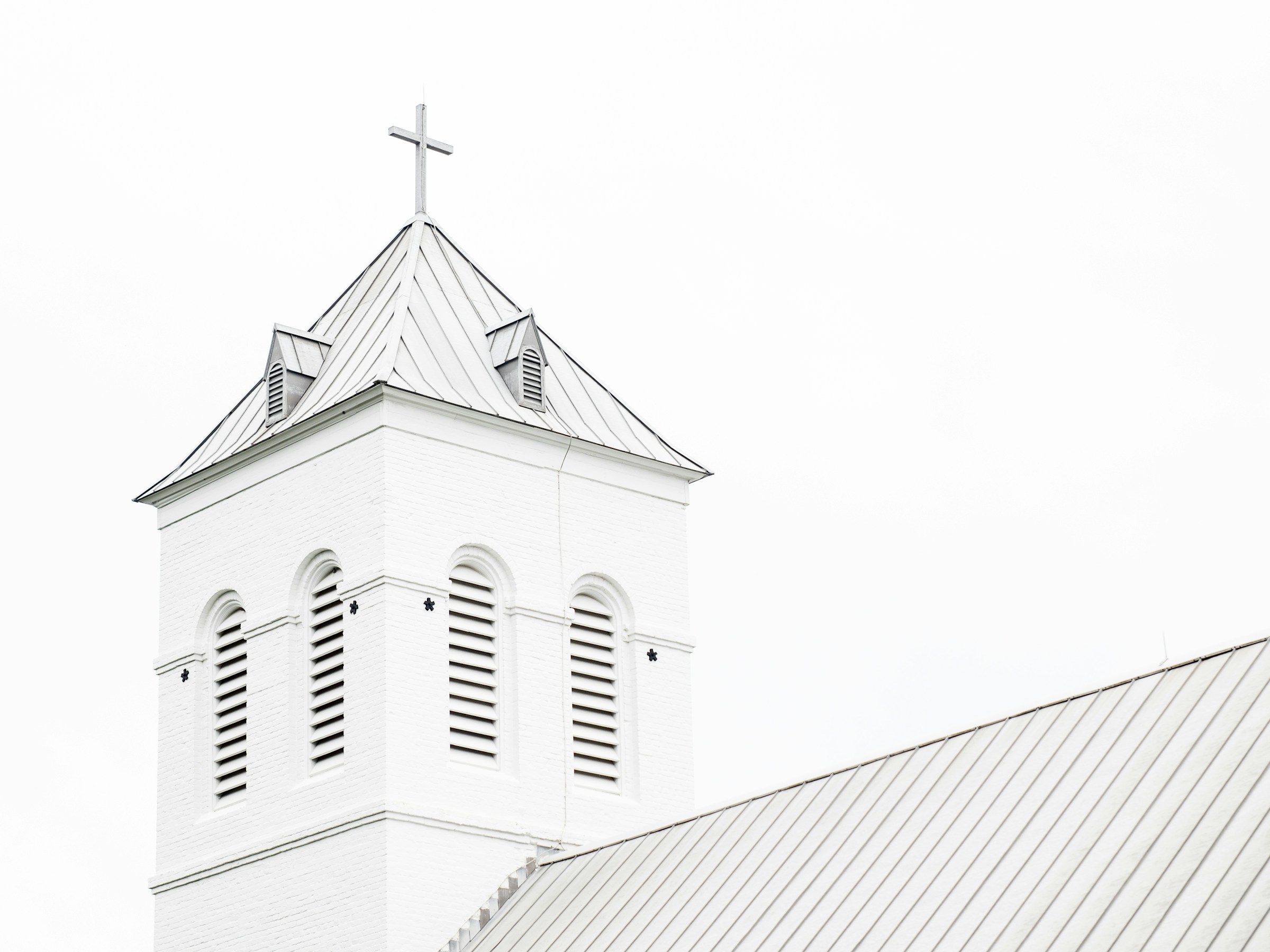 White church steeple with a cross atop a slanted roof, showcasing architectural details against a blank sky. Symbolizes faith and community.