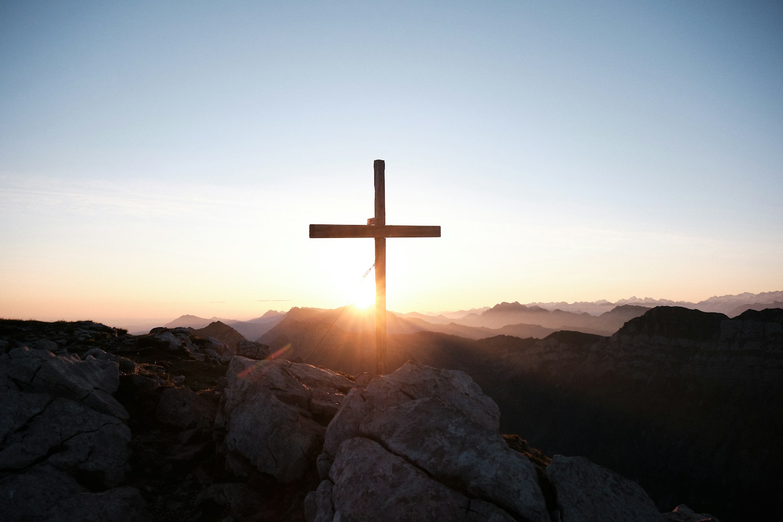 A wooden cross stands on a rocky peak, silhouetted against a vibrant sunrise over mountain ranges, symbolizing hope and reflection.