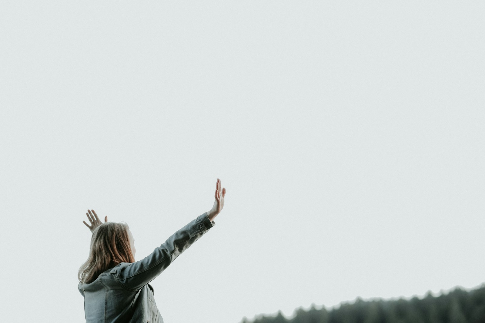 Person with long hair raises arms in a joyful pose against a pale sky, symbolizing freedom and connection with nature.