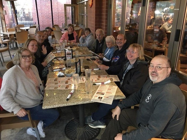A group of twelve people shares a meal at a restaurant, seated around a table with menus and drinks. They appear engaged and social.