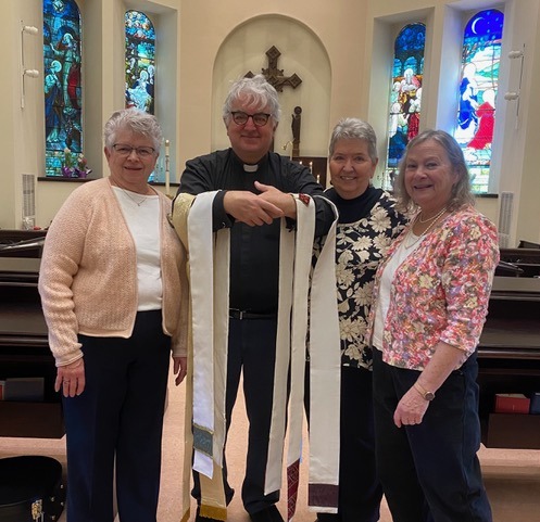 Four individuals stand together inside a church, with stained glass windows in the background. A man in the center holds ceremonial sashes, showcasing a religious event or celebration.