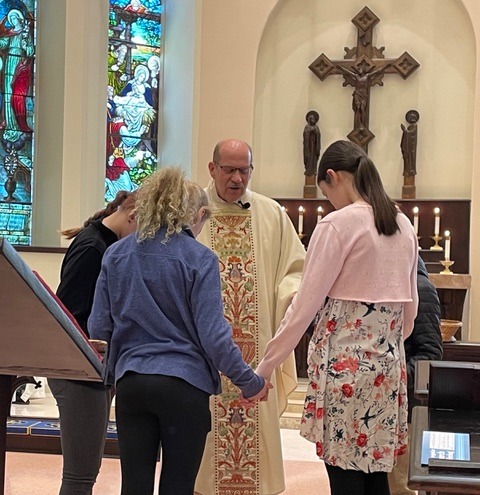 Clergy leads a group of children in prayer within a church, surrounded by stained glass and lit candles, emphasizing community and faith.
