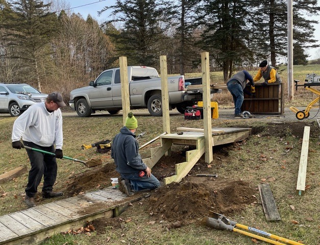 Workers construct a wooden ramp, digging and assembling alongside parked vehicles, highlighting community support for accessibility improvements.