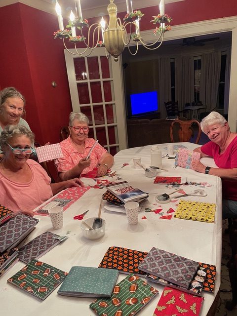 Four women craft at a table, creating decorated notebooks with colorful paper and scissors. A casual atmosphere highlights their friendship and creativity.