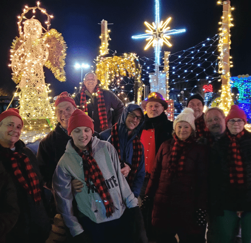A group of ten people in festive attire, including red hats and plaid scarves, smiles in front of brightly lit holiday decorations at night.
